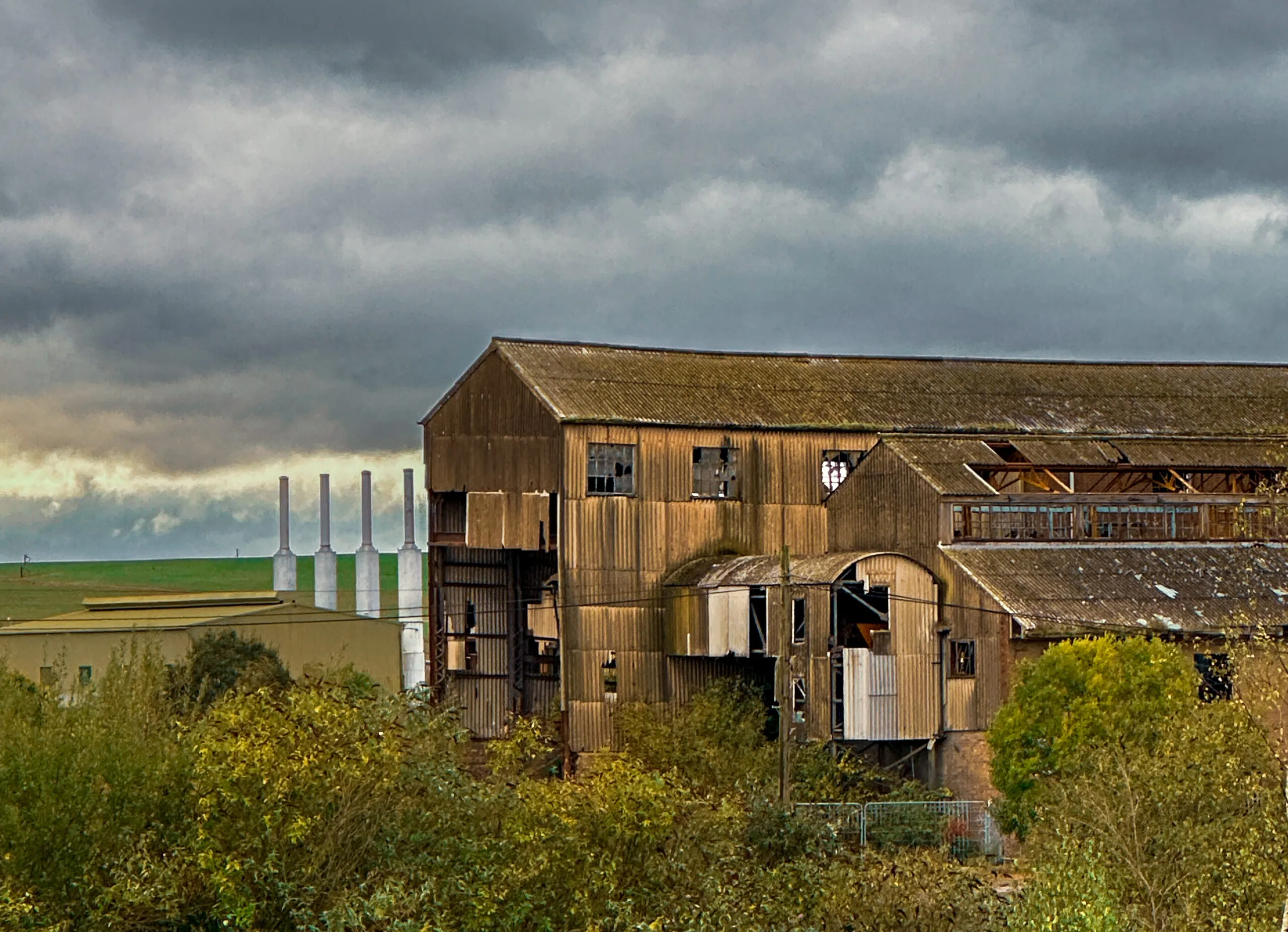 Corrugated-clad factory sheds with broken windows, four white kiln chimneys standing on the skyline behind